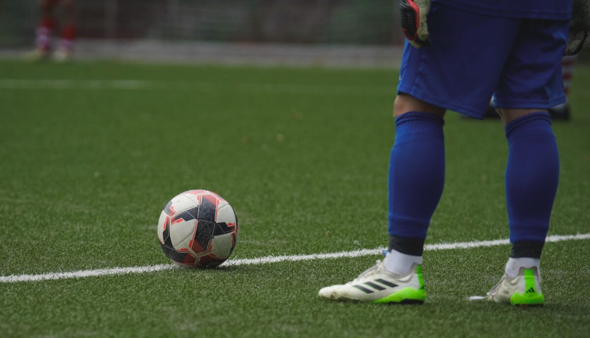 Close-up of a football player standing by a ball during a game on a grass pitch.